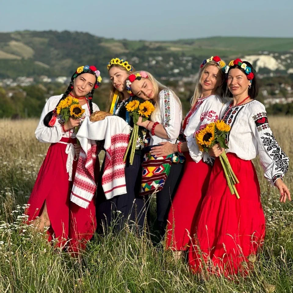 Four women dressed in traditional uUkrainian dress, carrying sunflowers and in a cosy friendly pose in a grassy field. It is a sunny day, there is a blue sky and there are out of focus hills in the background.